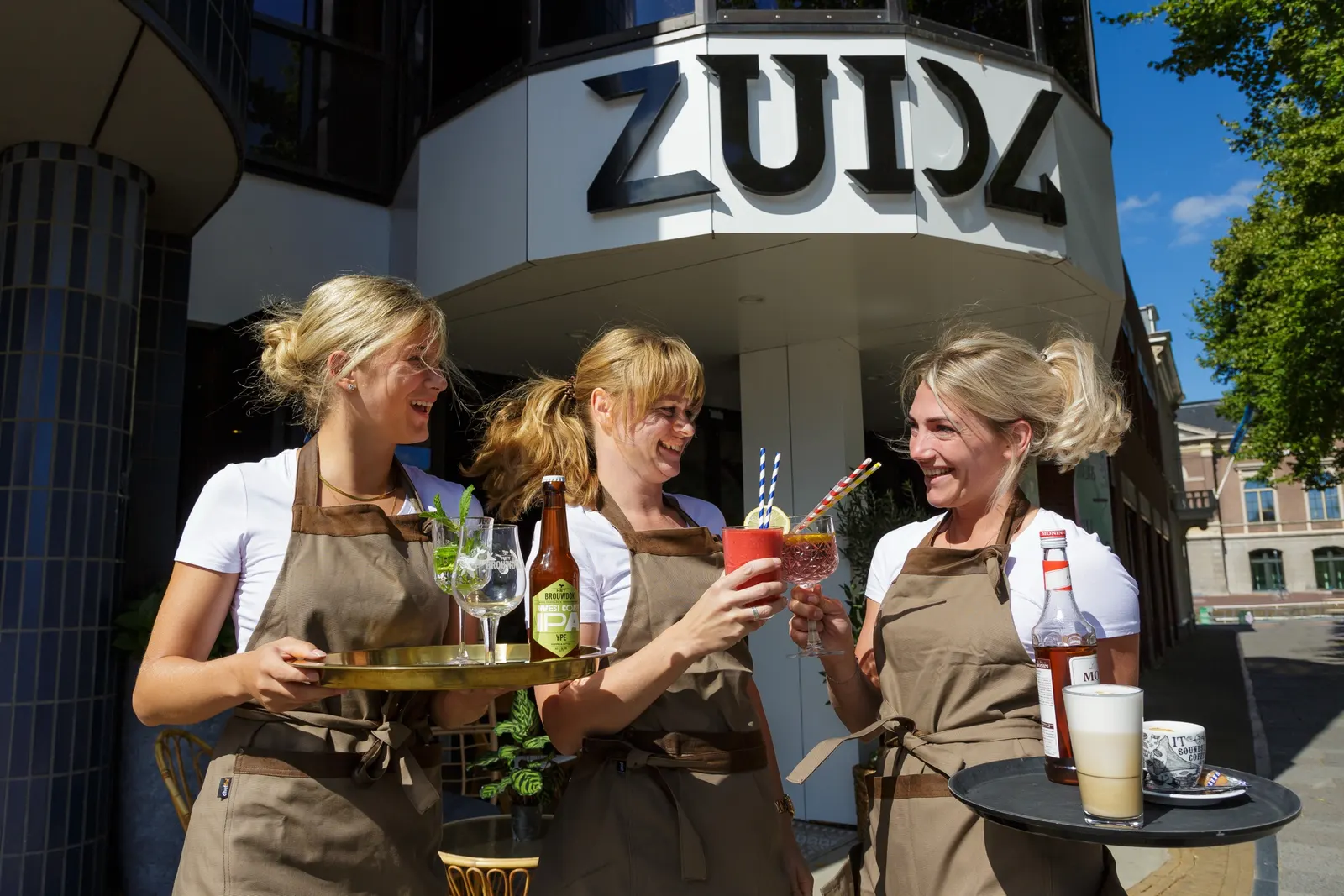 Drie medewerkers van Zuid 4 met dienbladen vol drankjes voor de ingang aan het Zuiderplein